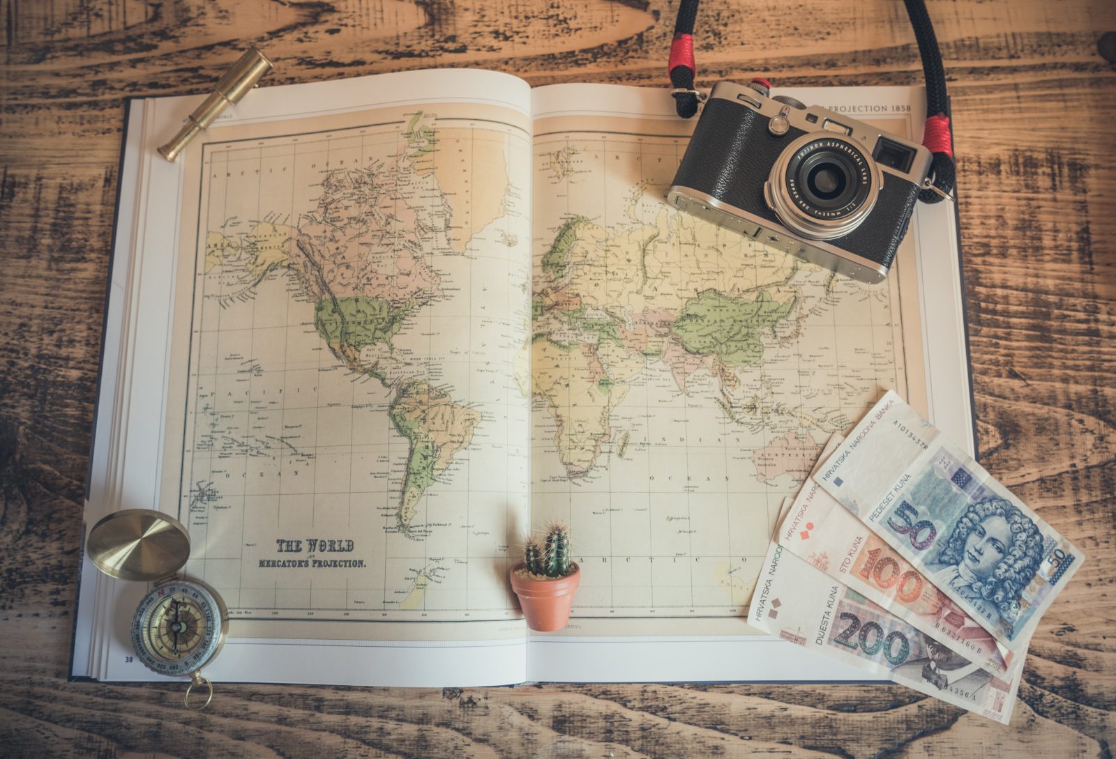 a map book on a wooden desk with a camera and money with a compass and tiny cactus, depicting travel.