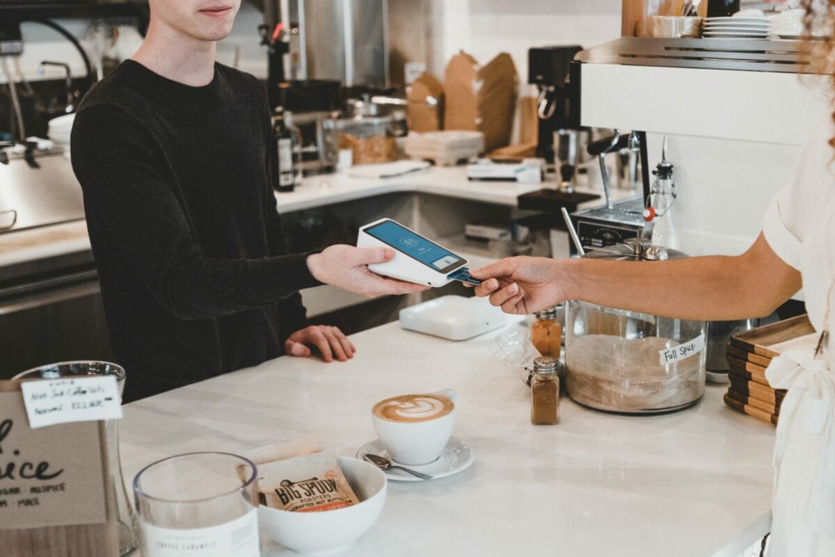 A man is handing a woman a payment machine at a coffee shop and she is putting her credit card into the reader.