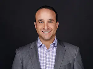 Dominick Vitolo, VP OF SECURITY SERVICES at MegaplanIT in a gray suit from the management team smiling at a black background.